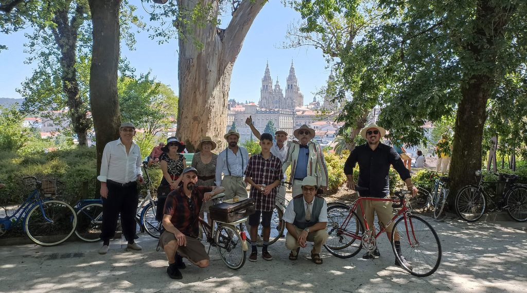 Participantes de la I Ruta de biciletas clásicas de Santiago posan a la sombra de los árboles con la catedral de fondo
